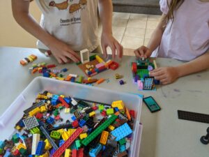 Two children build LEGO structures on a table, a shallow bin of LEGO pieces in the foreground.