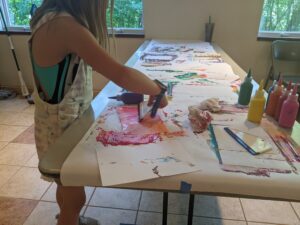 A child standing at a table using a handheld squeegee to manipulate paint on a piece of paper.