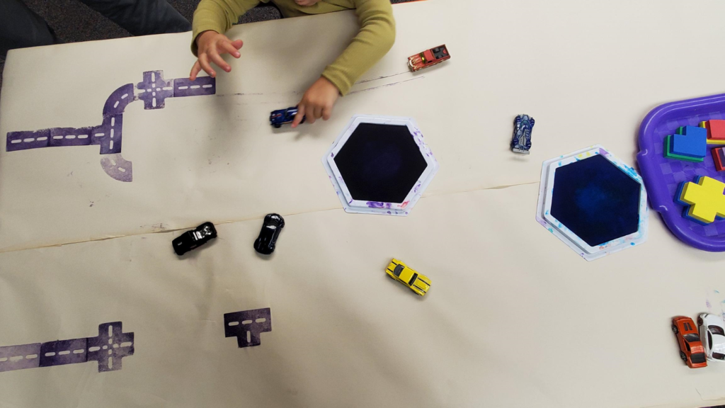 Top-down view of a table covered with butcher paper, on which a child plays with matchbox cars, plus large ink pads and stamps that leave a road-shaped pattern.