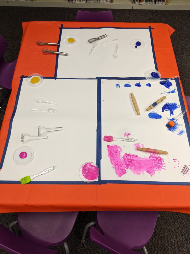 Photo of a table with taped-down pieces of paper, paint, and various utensils like plastic forks, rolling pins, and whisks.