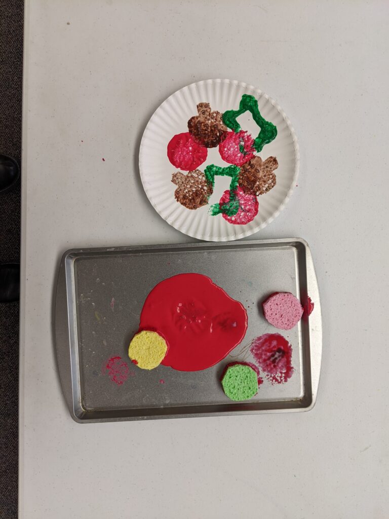 Photo of a baking tray with red paint and round sponges, and a paper plate covered in sponge paint prints to resemble pizza toppings.