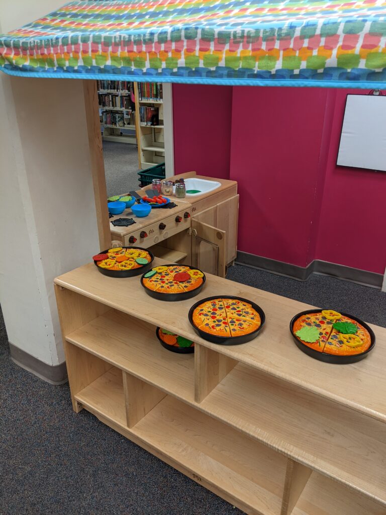 A wooden children's play kitchen, with plastic pizza and kitchen items upon the countertops.