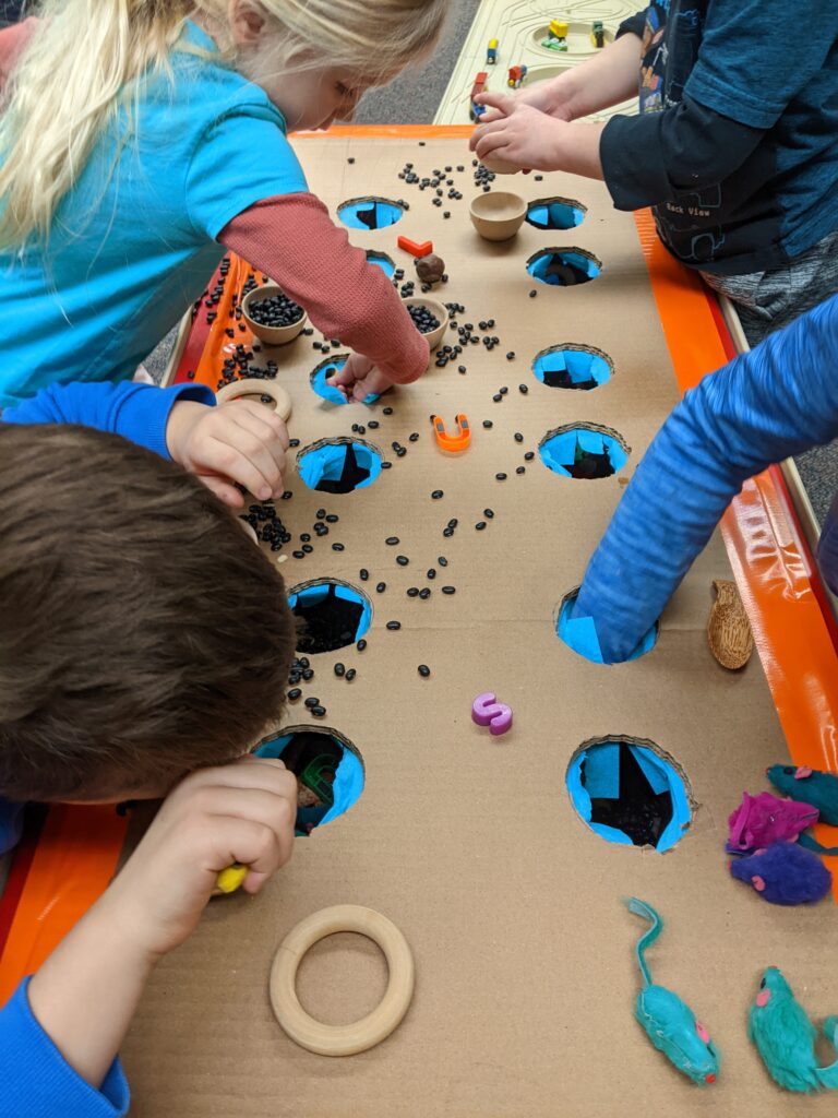 Close-up photo of children reaching their arms through holes cut in a cardboard sheet. On top of the cardboard are various items like black beans, wooden rings, and toy mice.