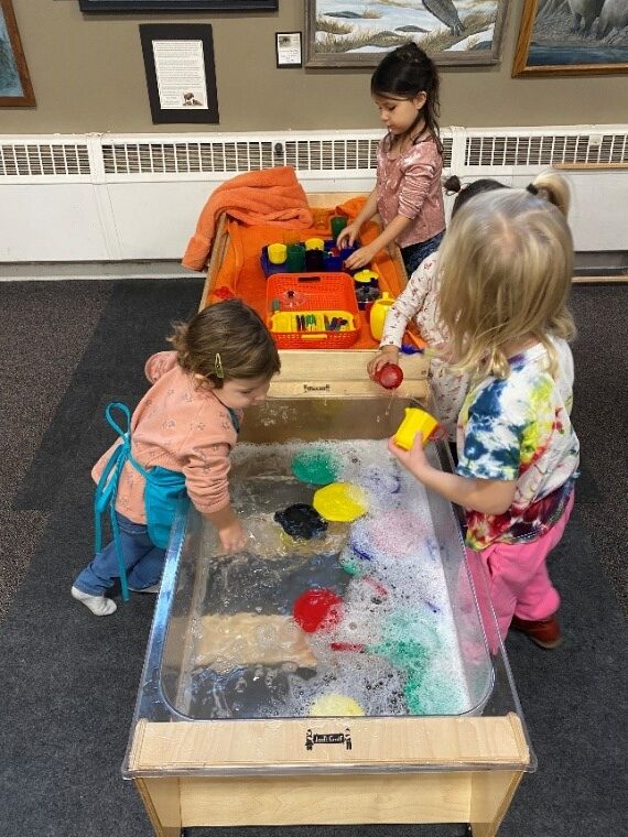 Photo of children playing in a tub of soapy water, "washing" toy plastic dishes. A second tub full of towels is the drying station.