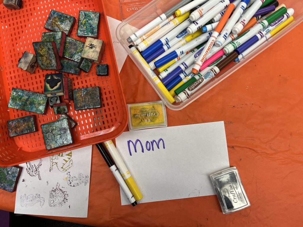 Photo of a tabetop with a tub of colored markers, a tub of dinosaur rubber stamps, and a paper with "Mom" written on it.