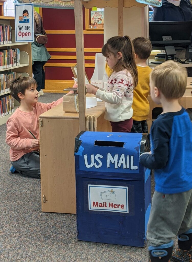 Photo of children playing at a wooden counter. One child holds an envelope. In the foreground is a blue cardboard box with "US MAIL" written on it.