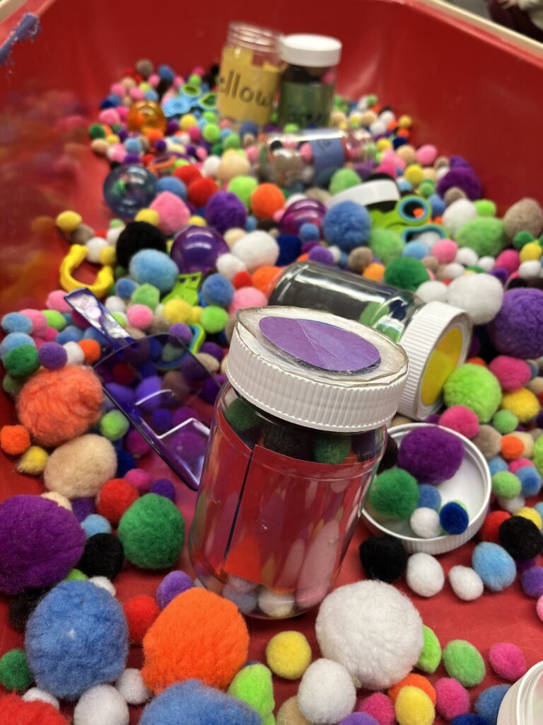 Close up photo of a red tub filled with various colored poms and clear plastic jars with colored labels.